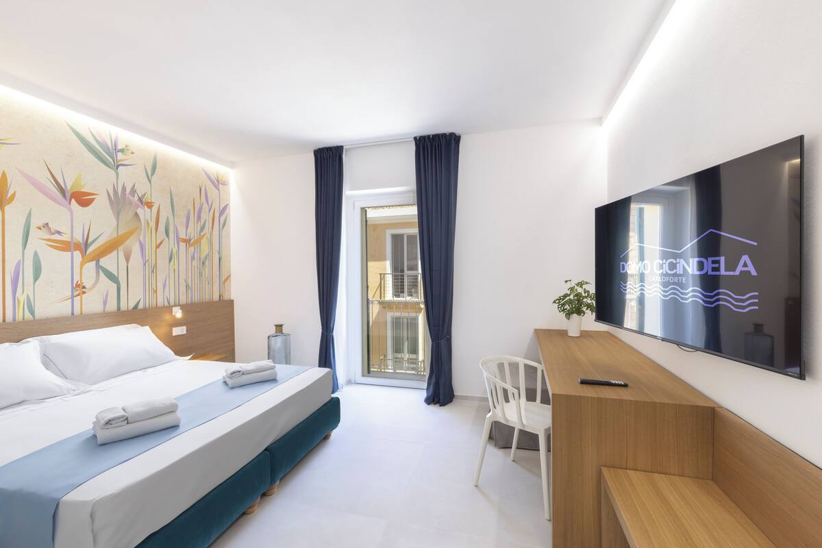 A modern hotel room with a bed, a desk, a chair, a TV, and a balcony with curtains.