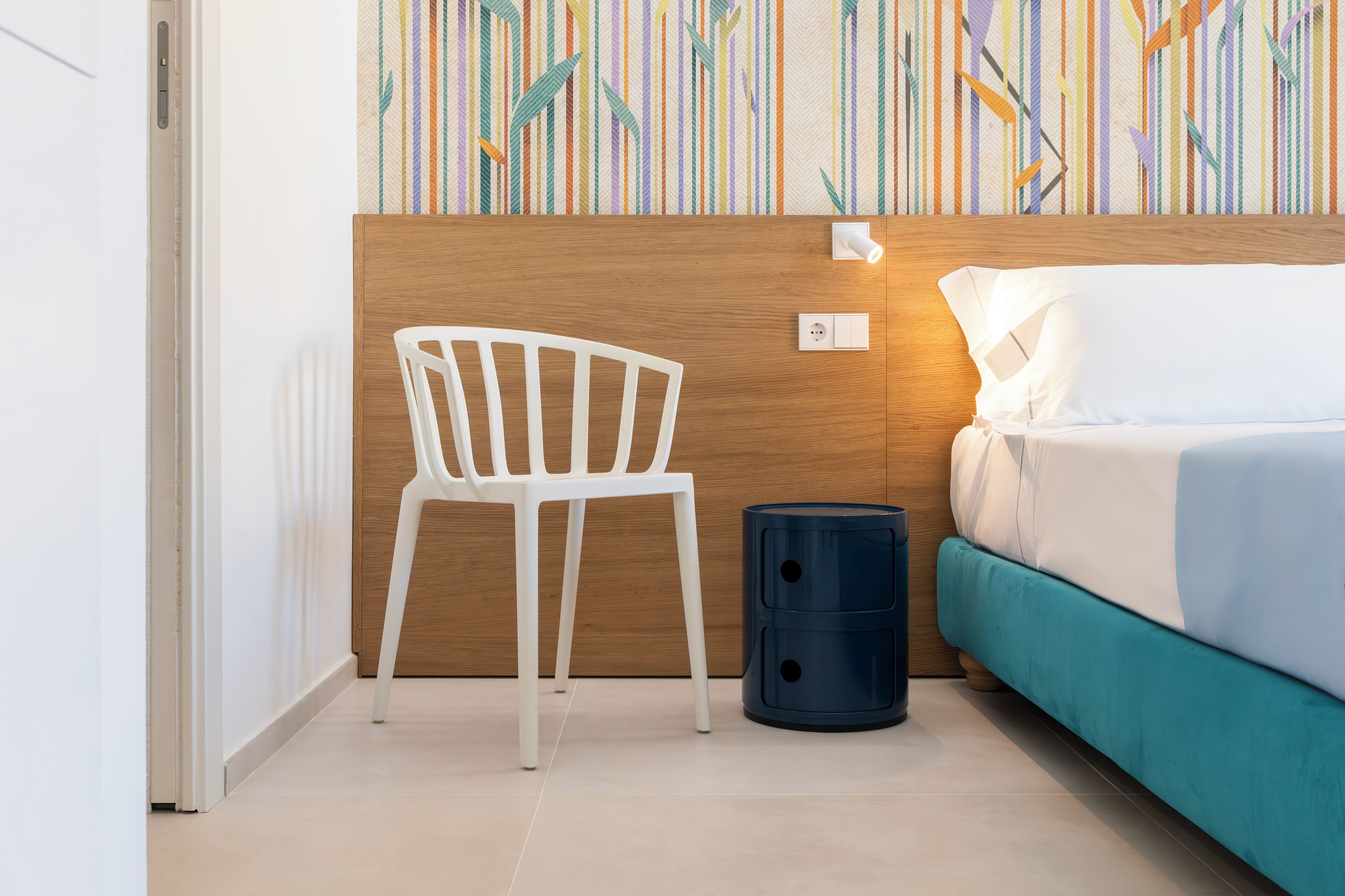 Een kamer met een houten hoofdeinde, een witte stoel, een blauwgroene bedbank en een blauwe prullenbak.