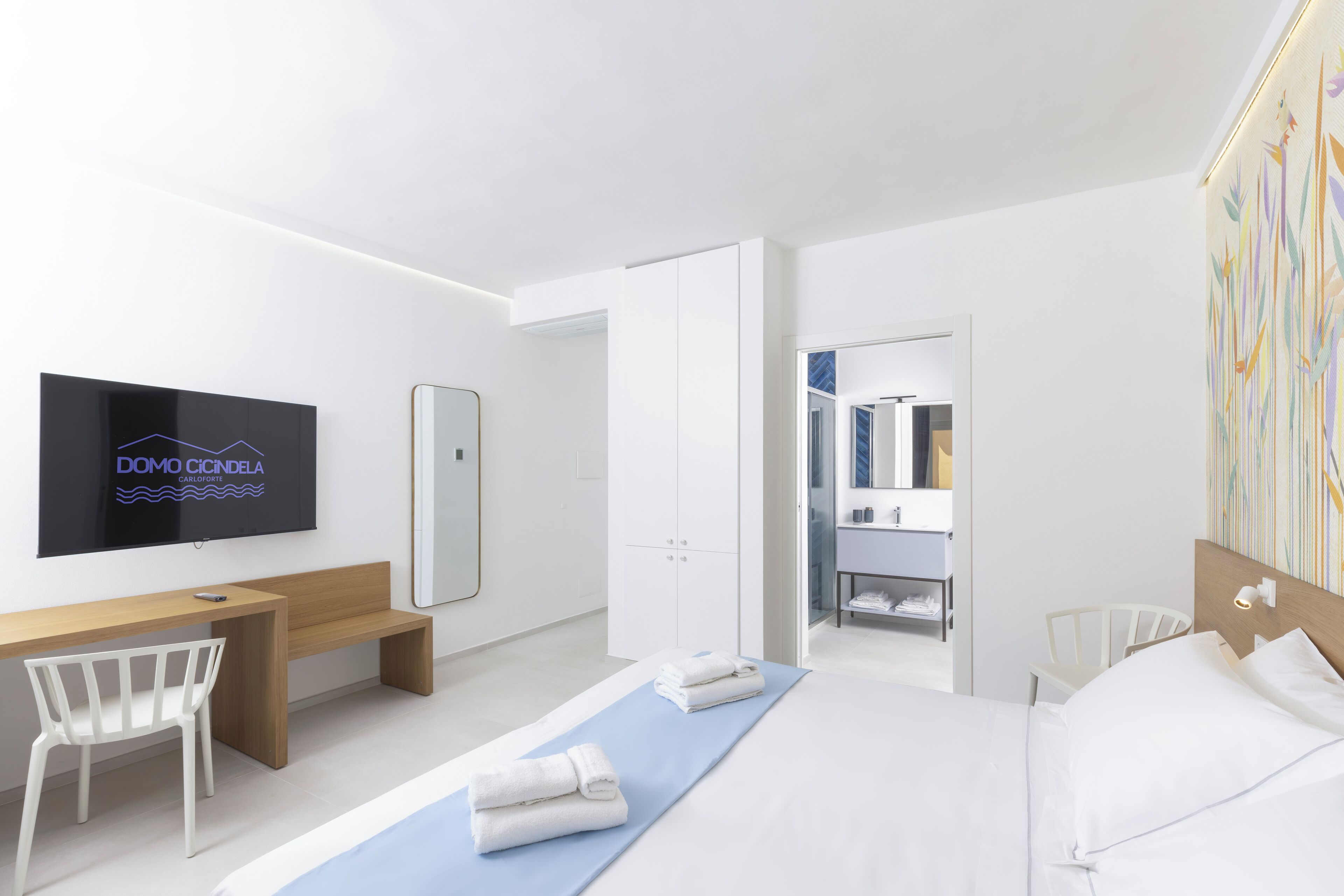 A modern hotel room with a large bed, a desk, a chair, a TV, and a bathroom visible through an open door.