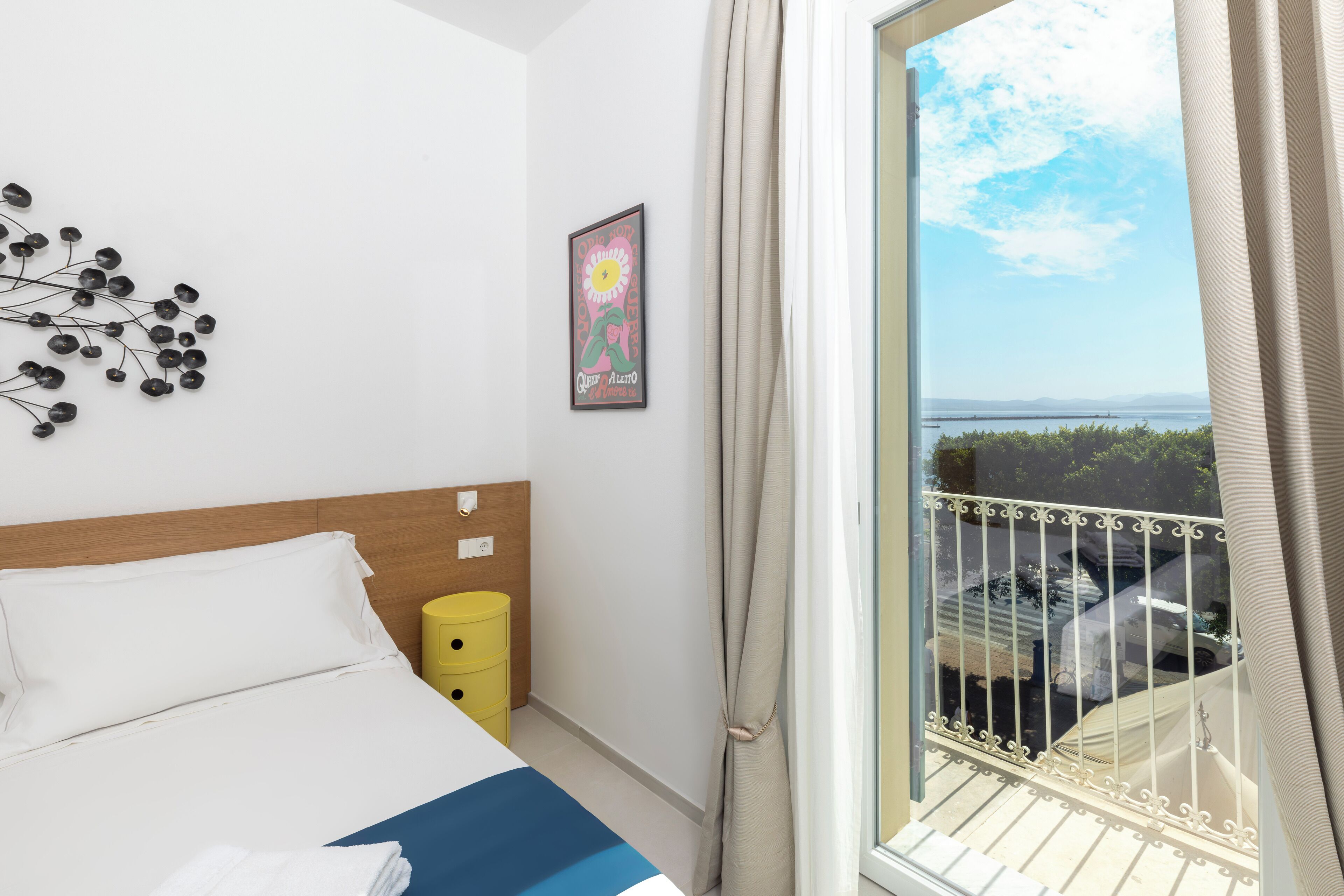 A hotel room with a bed, a bedside table, a yellow trash bin, a window with a view of the sea, and a decorative wall piece.
