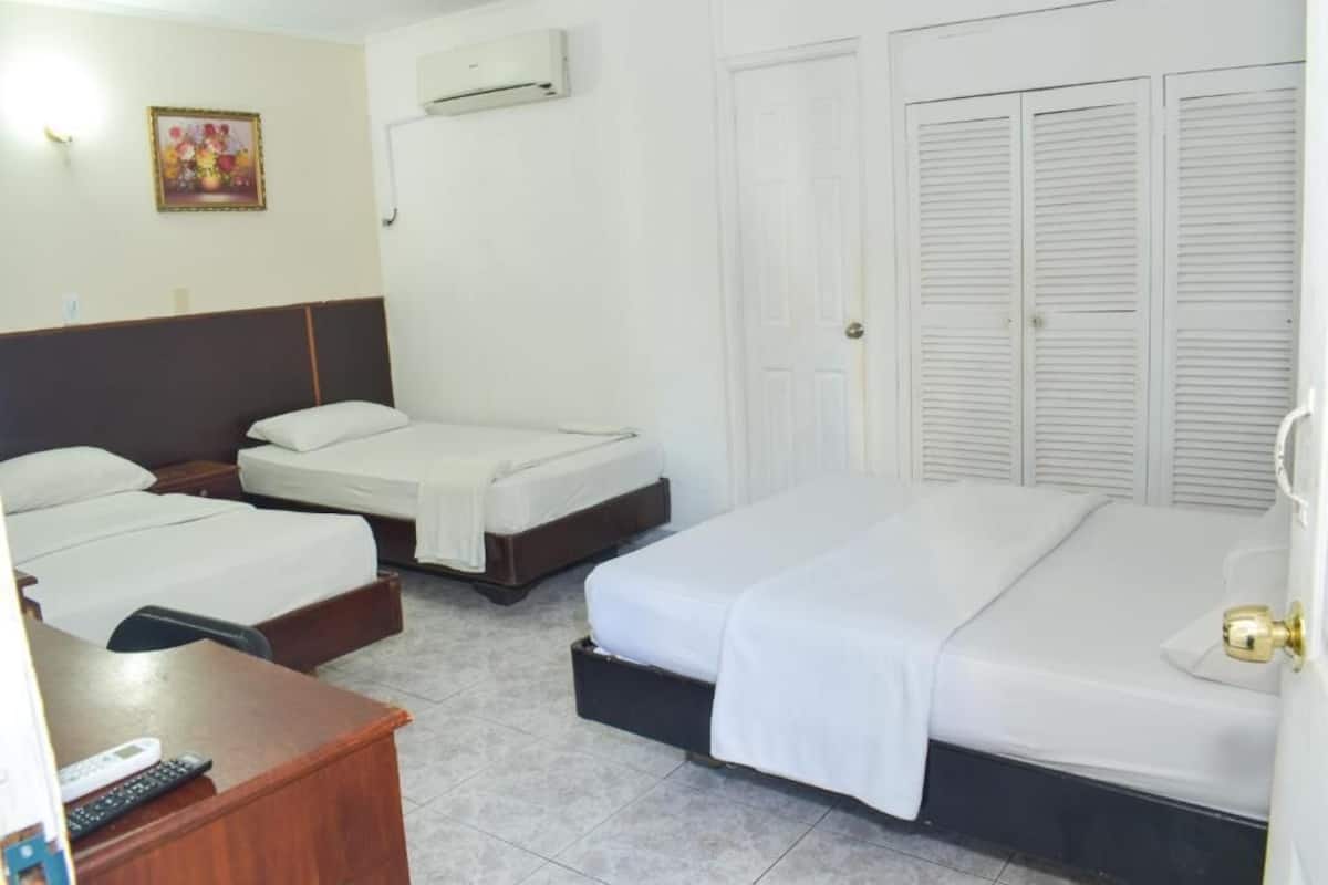 Une chambre d’hôtel avec deux lits simples, un grand lit, un bureau avec une télécommande, un climatiseur fixé au mur et une porte avec des volets à persiennes.