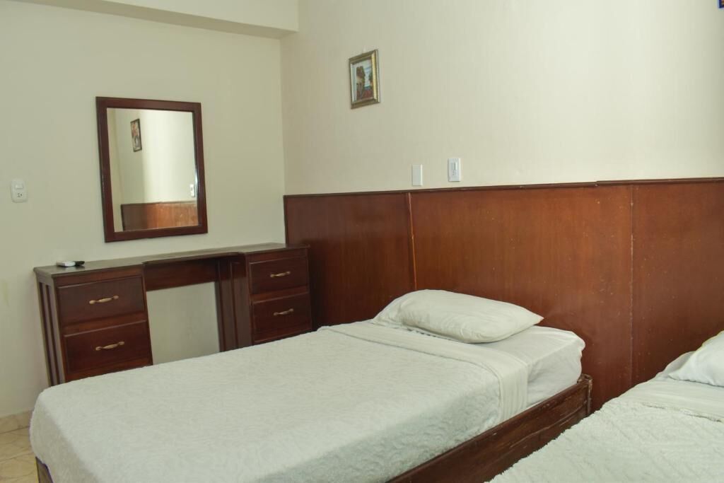 Een slaapkamer met een houten hoofdeinde, een bed met witte beddengoed, een dressoir met laden en een spiegel aan de muur.