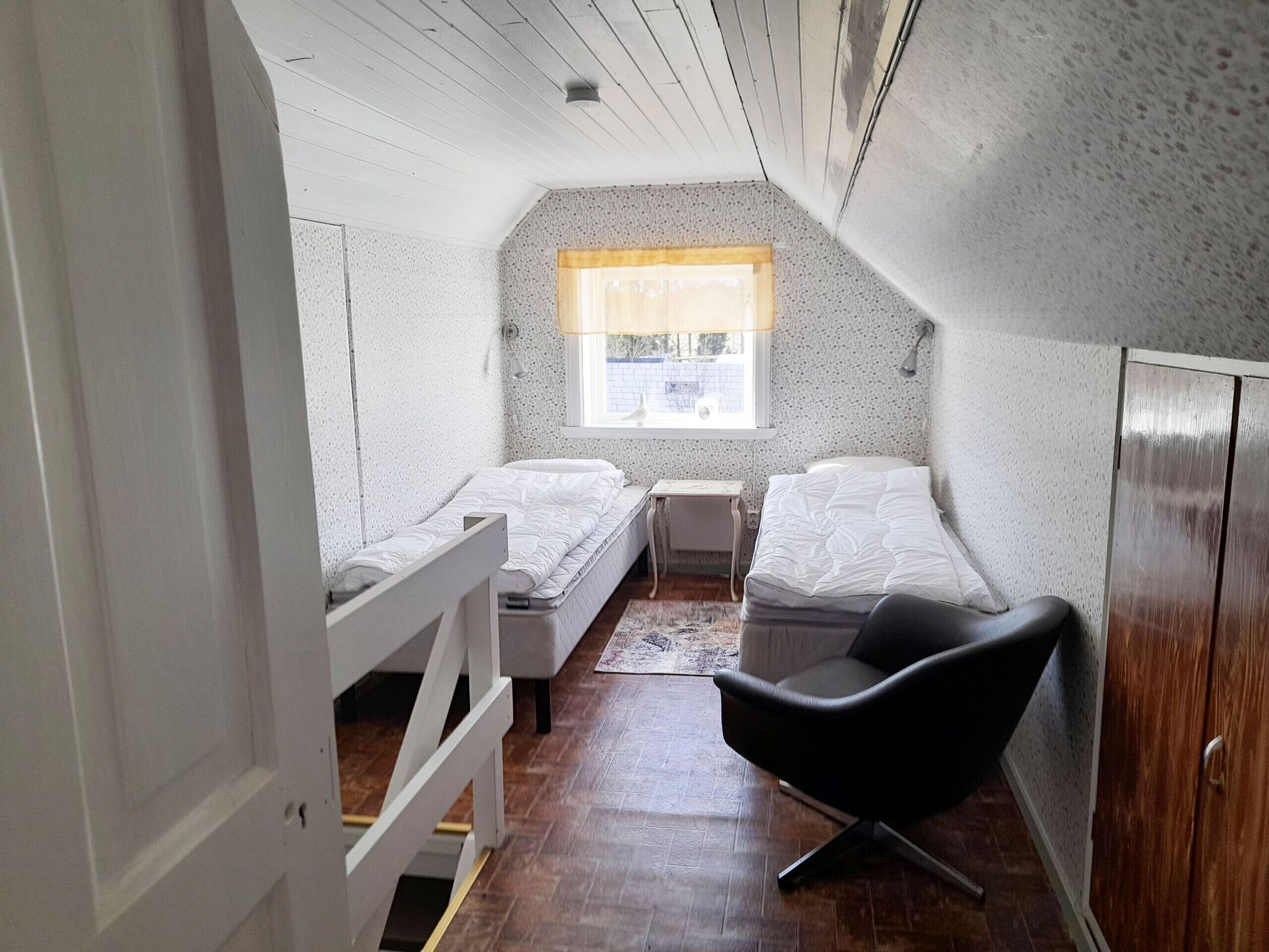 A small, cozy room with two single beds, a wooden wardrobe, a black chair, and a window with a view.