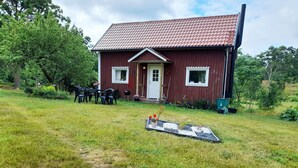 A red wooden house with a tiled roof, a front porch with chairs, and a garden area with a small pathway. - Cozy Country Cottage in Scenic Location near Mönsterås | SE05055 (Mönsterås)