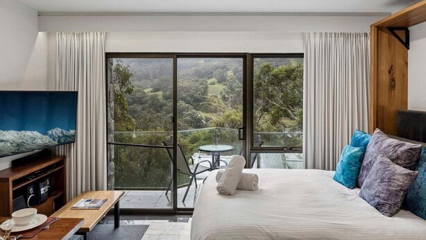 A bedroom with a large bed, a television, a dining table, and a balcony with a scenic view.