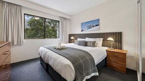 A bedroom with a large bed, a bedside table with lamps, a window with curtains, and a painting on the wall.
