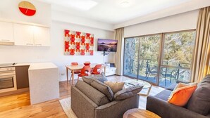 A modern living room with a sofa, a coffee table, and a TV. There is a kitchen area with cabinets and a dining table with chairs. Large windows provide a view of the outdoors. - Lantern 07 - Balcony (Thredbo)