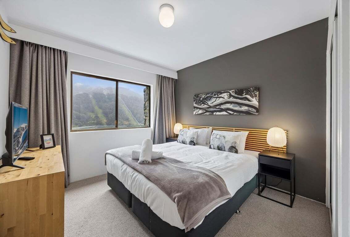 A modern bedroom with a large bed, a desk, a nightstand, and a window with a mountain view.