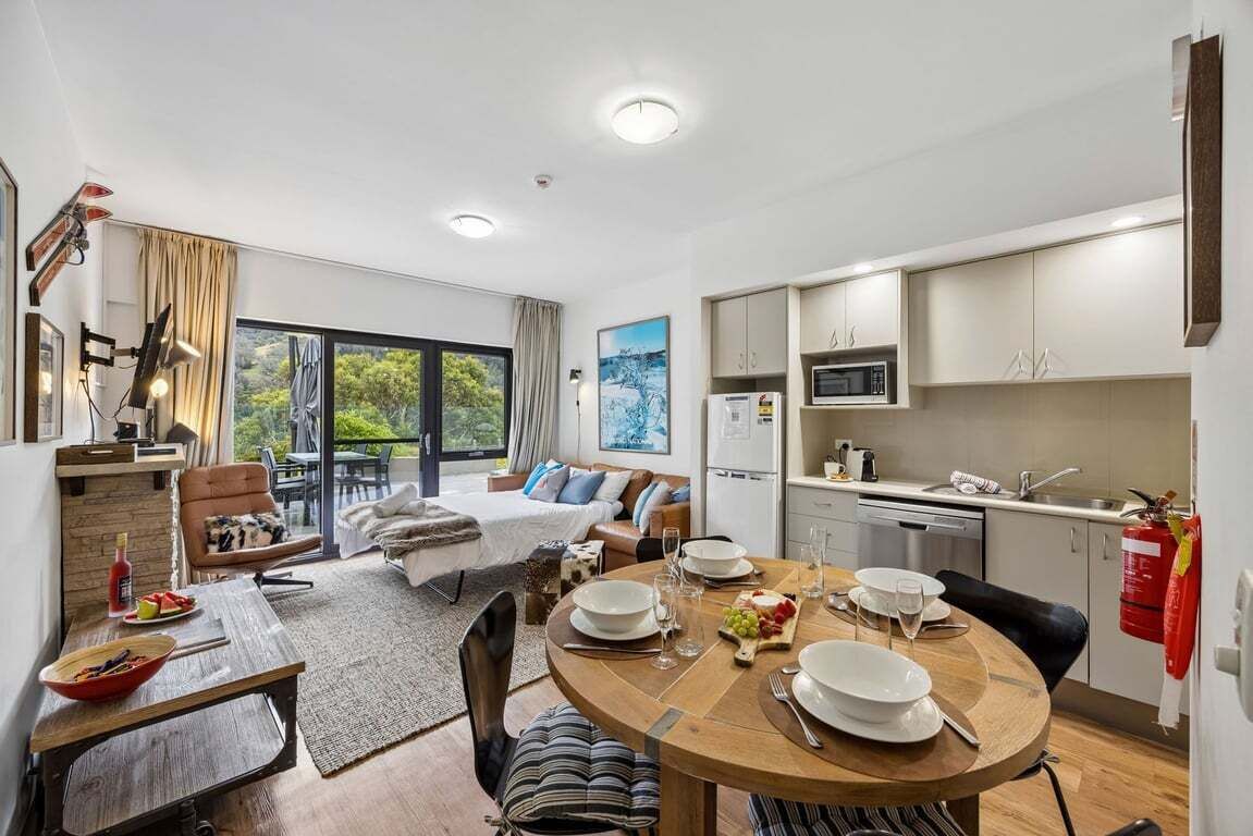 A modern, well-lit room with a dining table set for four, a bed with a view, and a kitchenette.