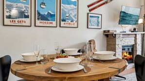 A dining area with a round wooden table set for four, three framed posters on the wall, a fireplace, and a television. - Lantern 14 - Terrace (Thredbo)