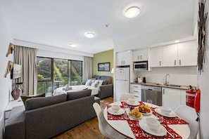 A modern living room with a sofa, dining table set for two, and a kitchen area with white cabinets and appliances.