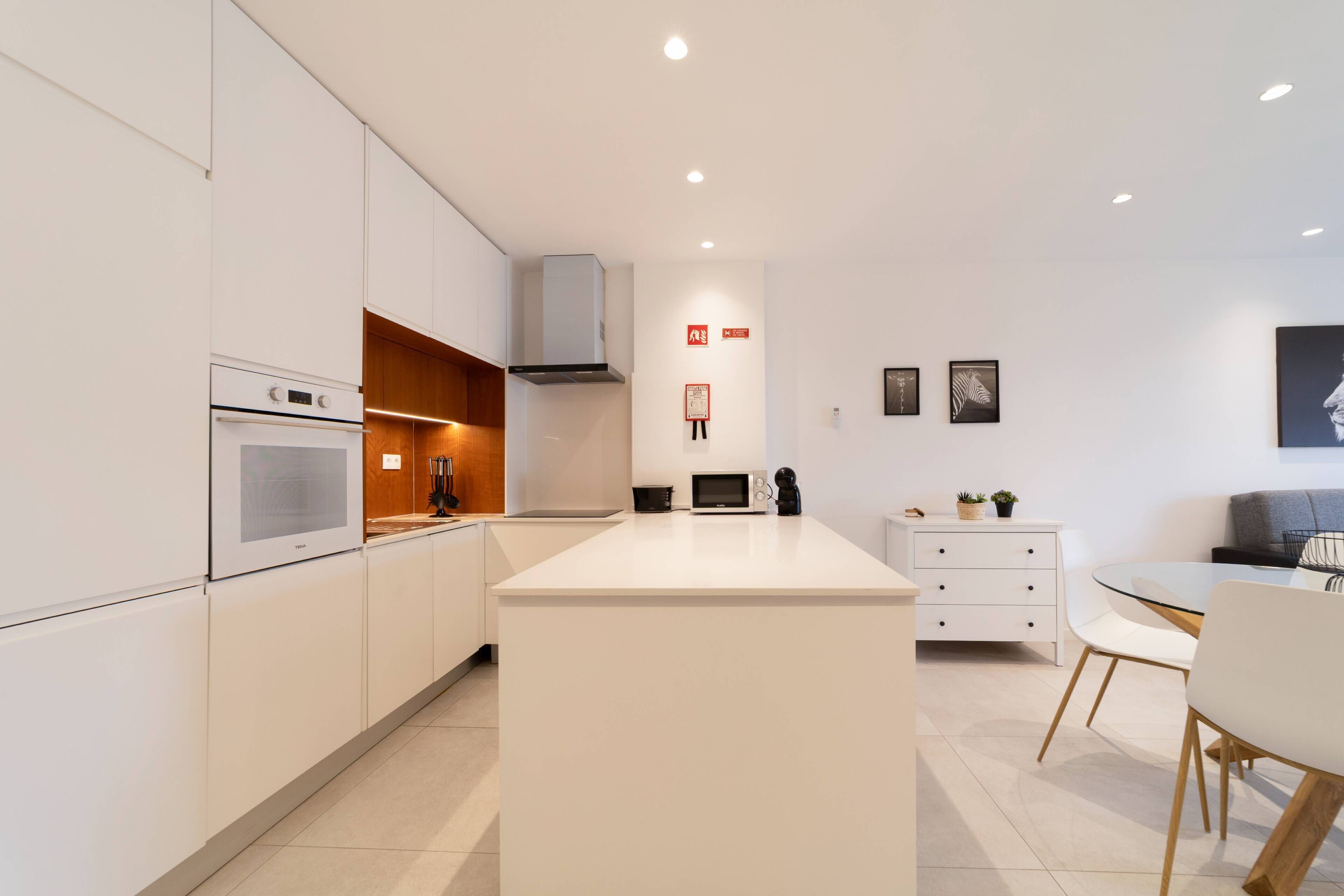 Una cocina moderna con armarios blancos, una isla central y un comedor con mesa y sillas de vidrio.