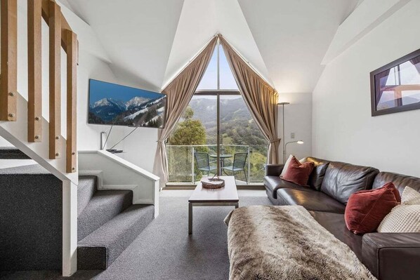 A modern living room with a staircase, a large window with curtains, a leather sofa, and a flat-screen TV.