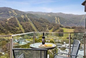 A balcony with a table, a bottle of wine, and glasses, overlooking a mountainous landscape.