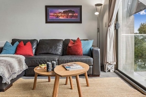 A modern living room with a leather sofa, colorful cushions, a round wooden coffee table, and a framed picture on the wall.