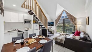 A modern kitchen with white cabinets, a wooden staircase, a dining area with a table set for two, and a sofa with a view of the outdoors.