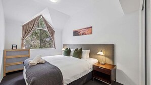 A bedroom with a bed, bedside table, lamp, window with curtains, and a picture on the wall.