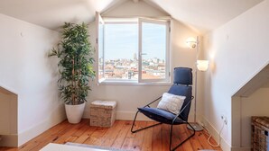 Une pièce chaleureuse avec un plancher en bois, une chaise noire avec un coussin, une grande plante en pot et une vue sur la ville par la fenêtre.