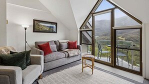 A modern loft bedroom with a sloped ceiling, a large window offering a view of the countryside, a grey sofa with red cushions, and a small wooden stool. - Lantern 30 - Balcony (Thredbo)