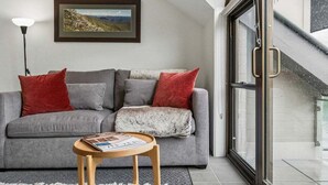 A living room with a grey sofa, red and white cushions, a small wooden side table with a magazine, a floor lamp, and a framed picture on the wall. - Lantern 30 - Balcony (Thredbo)