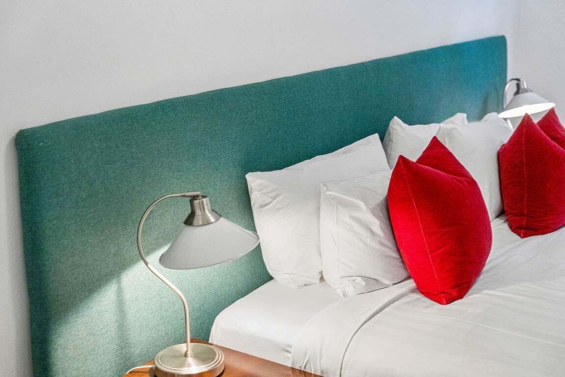 A bed with white bedding and red pillows, a teal headboard, and a bedside lamp.