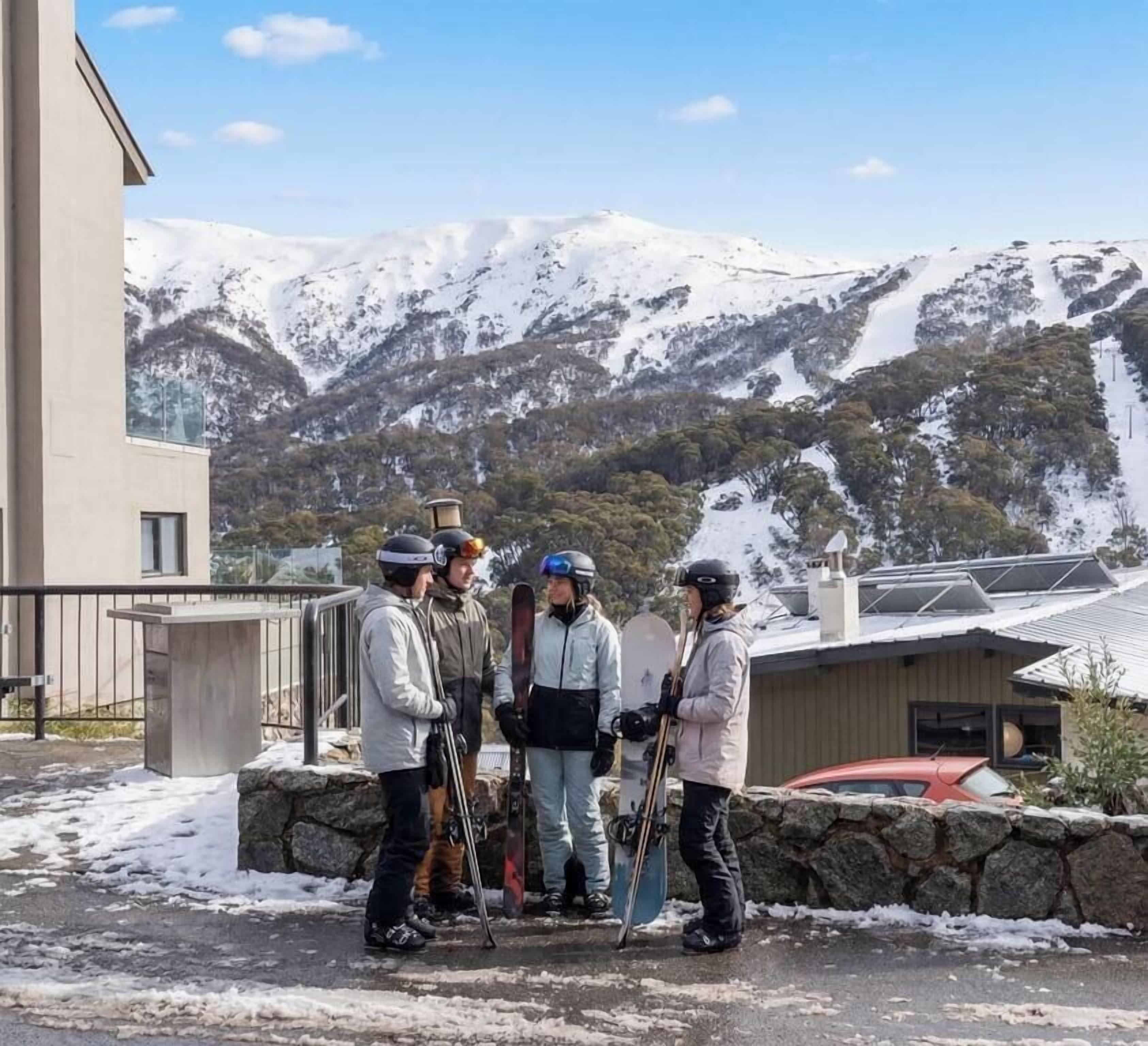 Un grupo de personas con esquíes frente a un edificio con vistas a montañas nevadas.