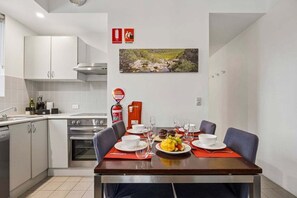 A modern kitchen with white cabinets, a stove, and a dining area with a table set for two. There are fire safety signs on the wall and a painting above the dining table.