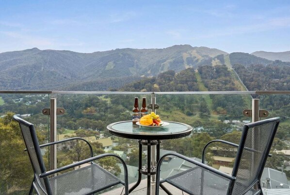 A balcony with two chairs, a table with food and drinks, and a scenic mountain view. - Lantern 33 - Balcony (Thredbo)