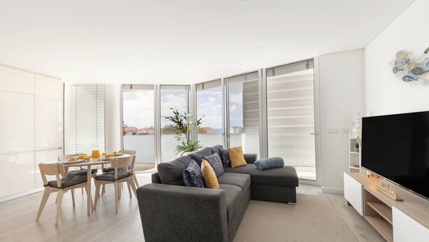 A modern living room with a grey sofa, a flat-screen TV, and a dining area with a table and chairs. - Atlantic Pearl – 5 min to the beach w/ parking (Nazaré)