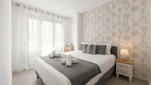 A neatly made bed with a grey throw blanket, a bedside table with a lamp, and a floral wallpaper pattern on the wall. - Atlantic Pearl – 5 min to the beach w/ parking (Nazaré)