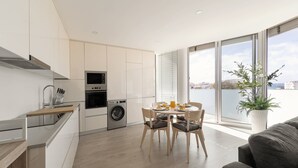 A modern kitchen with a washing machine, a dining area with a table and chairs, and a large window with a view of the sea. - Atlantic Pearl – 5 min to the beach w/ parking (Nazaré)