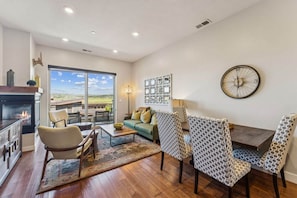 A spacious living room with a fireplace, a large window offering a view of the outdoors, a dining area with patterned chairs, and a wall clock. - Park Lane South Departments (Park City)