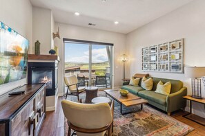 A spacious living room with a fireplace, comfortable seating, a large window with a view, and a wall-mounted display. - Park Lane South Departments (Park City)