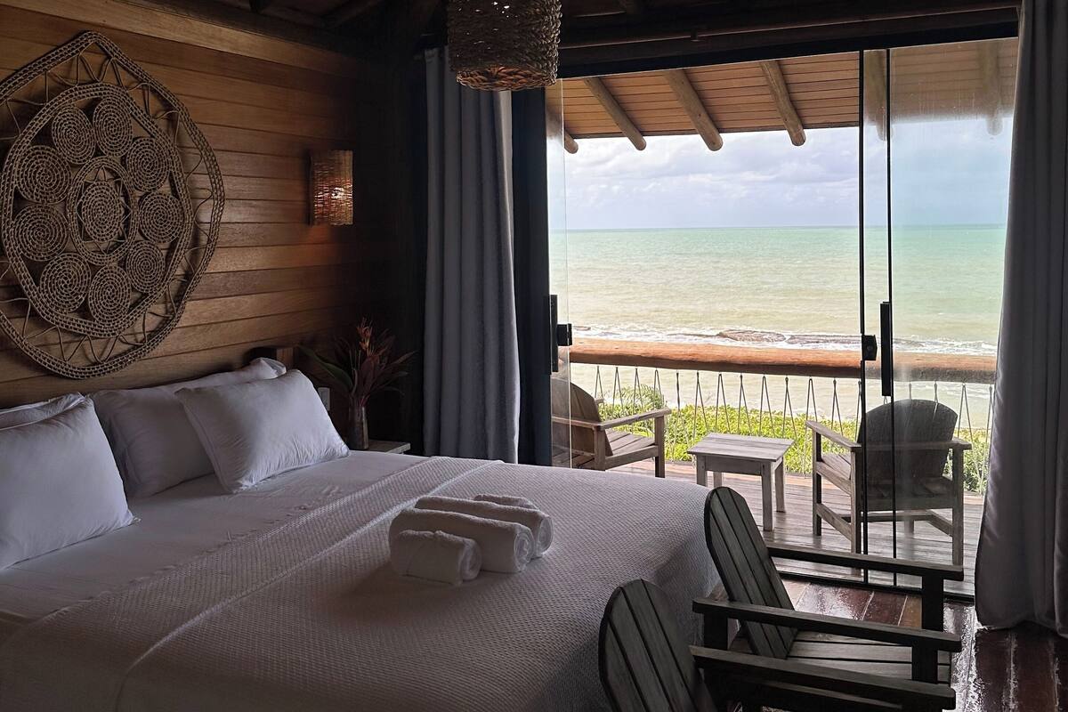 A bedroom with a large bed, a view of the ocean, wooden ceiling beams, and a woven wall hanging.