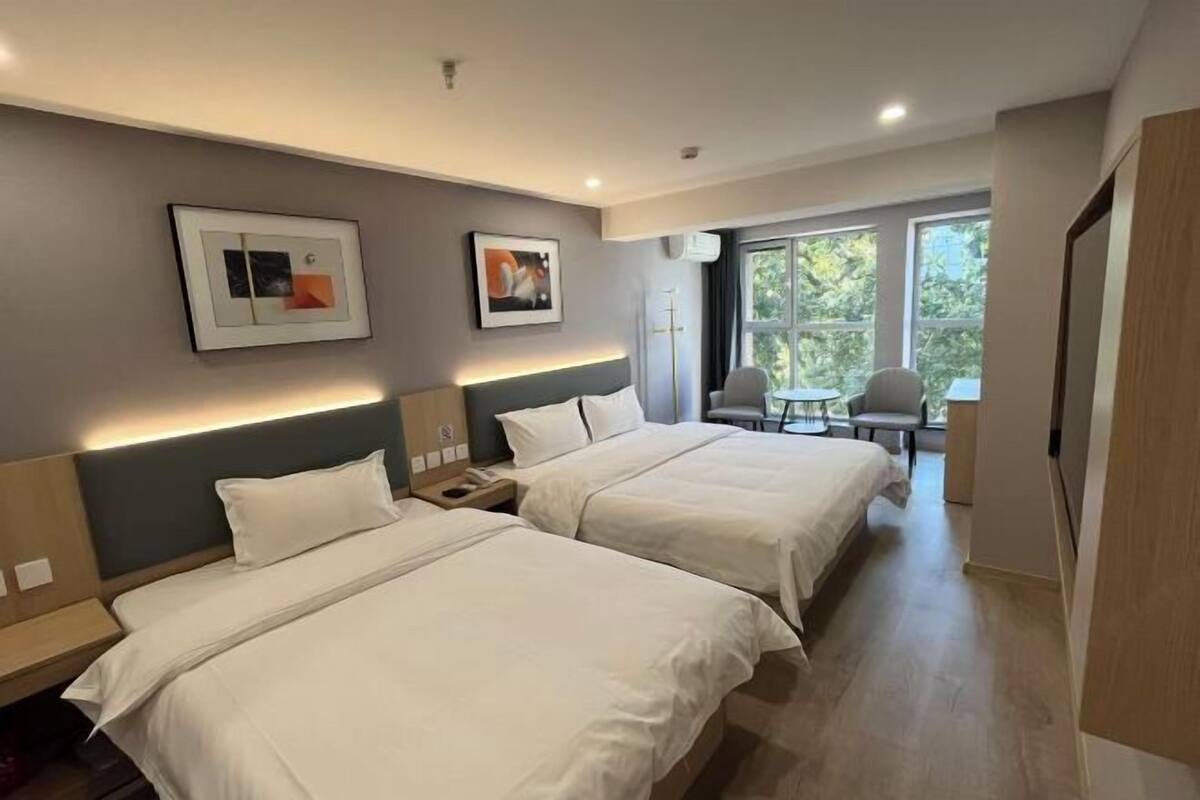 Une chambre d’hôtel avec deux lits, un bureau, une chaise et une fenêtre donnant sur des arbres.