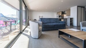 Een moderne woonkamer met een bank, fauteuil en salontafel, een balkon met kleurrijke stoelen, en een keukenhoek.