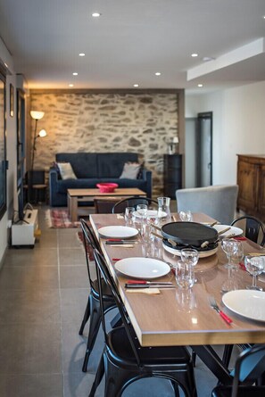 A dining area with a long table set for four, a stone wall in the background, and a sofa in the adjacent living room.