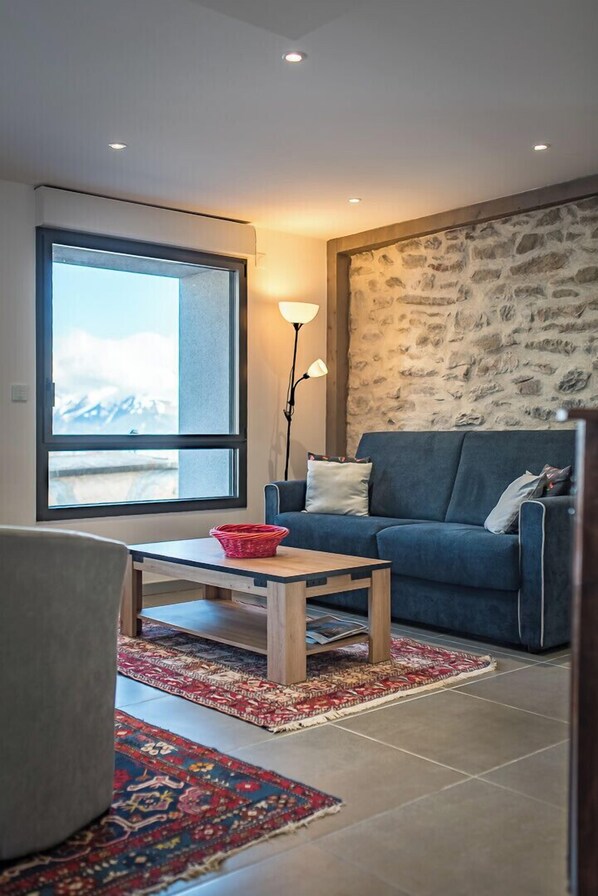 A living room with a large window, a blue sofa, a wooden coffee table, and a patterned rug.