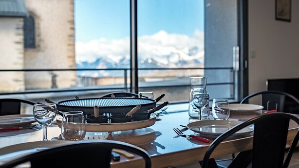 A dining table with a grill, glasses, and plates, set against a backdrop of a mountain view through large windows.