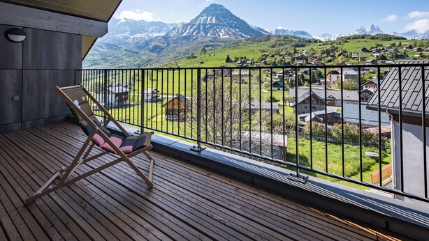 A wooden deck with a lounge chair, a balcony railing, and a view of a mountain range and a village below.