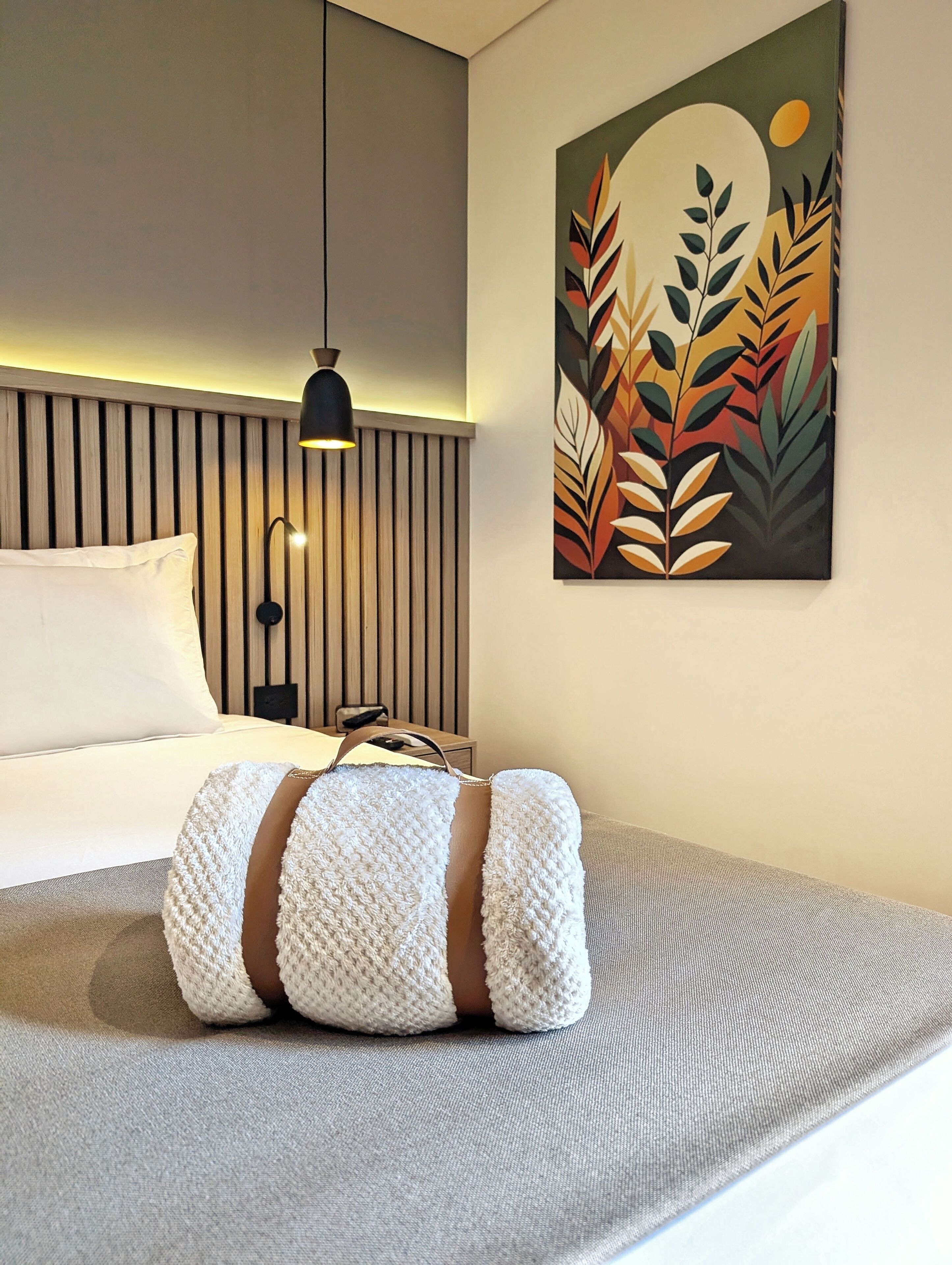 A modern hotel room with a bed, a wall-mounted lamp, and a painting of stylized leaves.
