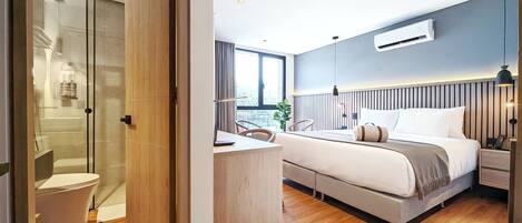Una habitación de hotel moderna con suelo de madera, cama con ropa blanca, escritorio con silla, cabina de ducha de vidrio y vistas al exterior.