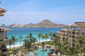 A resort with a pool, palm trees, and a view of the ocean and mountains. - Ocean-view Cabo villa w/ terrace, BBQ, hot tub — steps from Medano Beach!  #1607 (Cabo San Lucas)