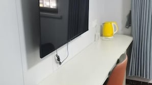 Ett rum med en platt-TV som är monterad på väggen, ett vitt skrivbord, en gul mugg och ett fönster med gardiner.