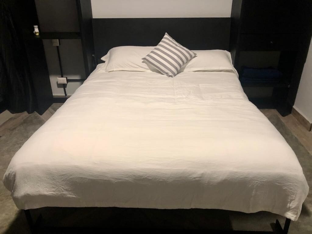 A neatly made bed with white linens and a striped pillow in a modern hotel room with dark wood headboard and a visible power outlet on the wall.