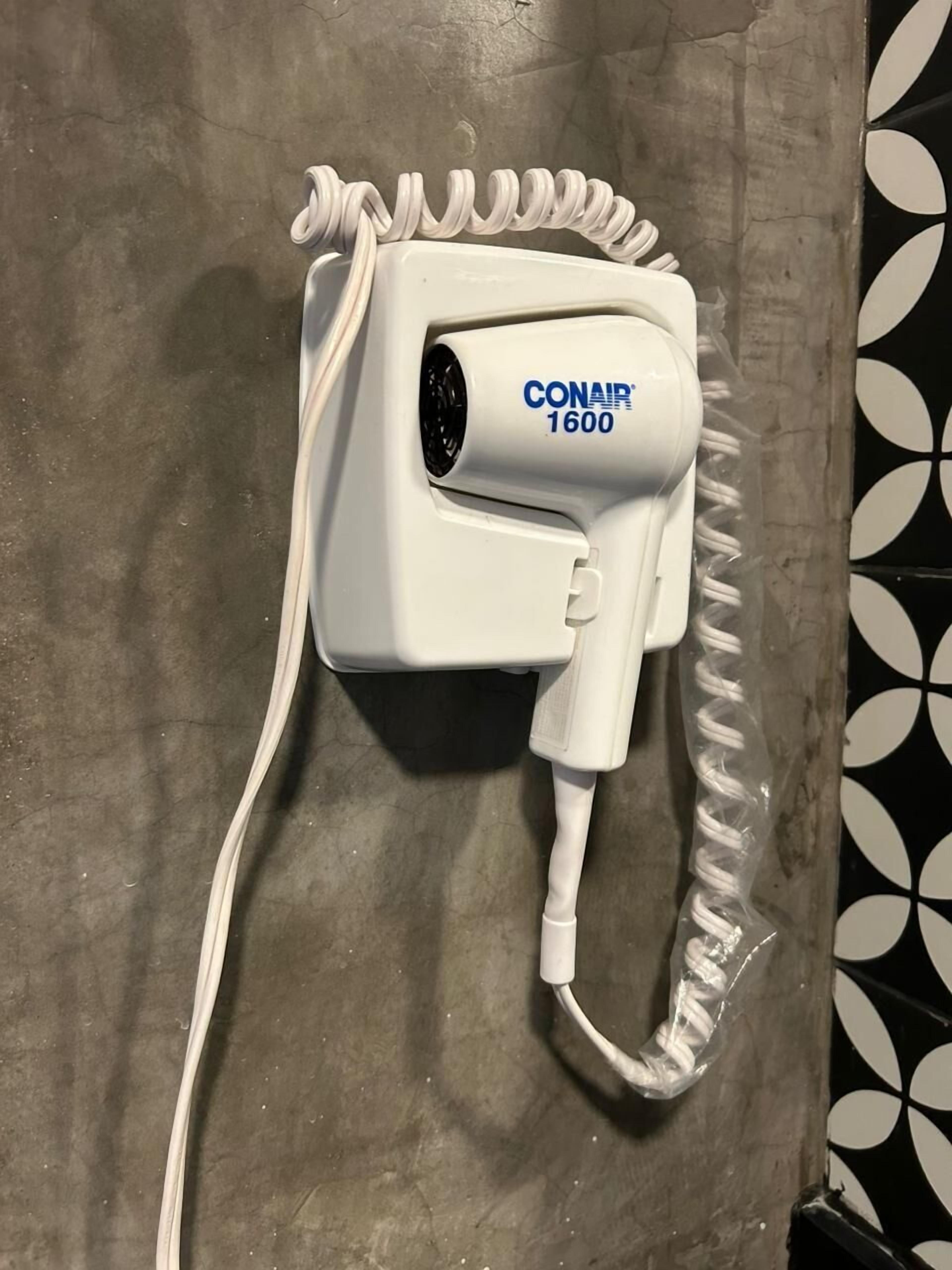 Secador de cabelo branco com fio, identificado como 'CONAIR 1600', fixado numa parede cinzenta.