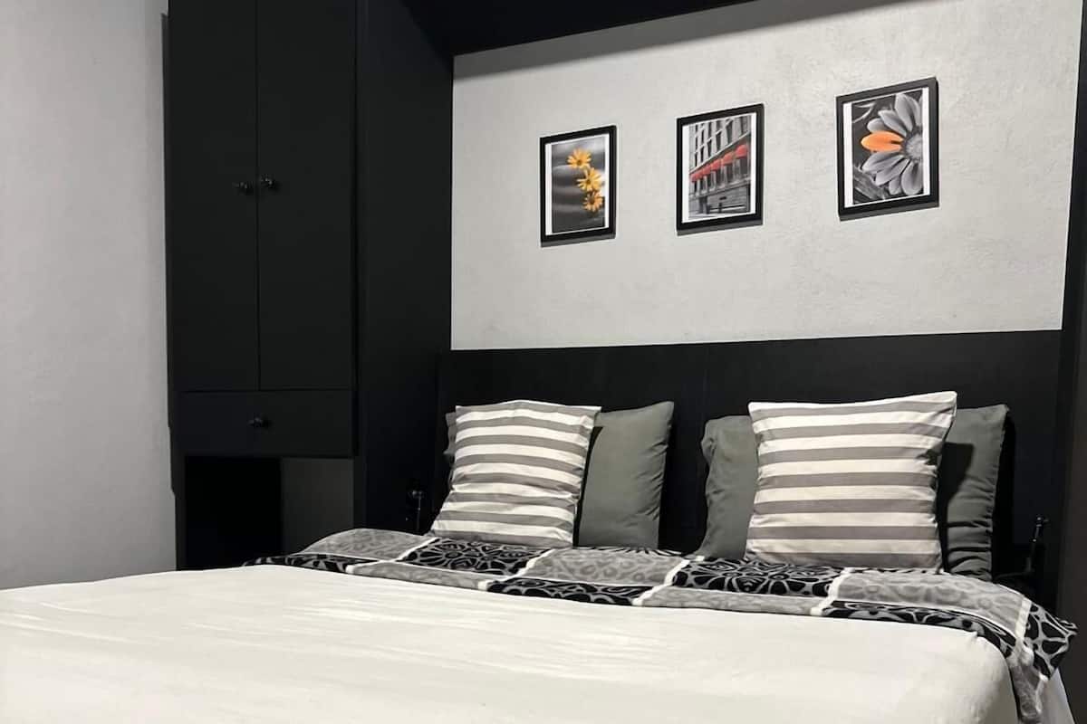 Ein ordentlich bezogenes Bett mit weißer Daunendecke und gestreiften Kissen, ein dunkler Kleiderschrank und drei gerahmte Bilder an der Wand.