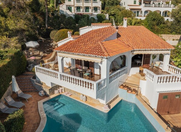 A villa with a swimming pool, a covered patio, and a view of surrounding buildings.