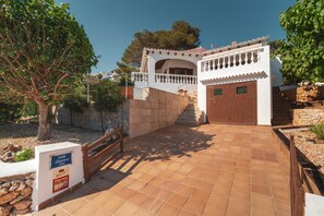 A villa entrance with a tiled pathway leading to a wooden door, a stone wall, and a sign indicating the villa name and room number.