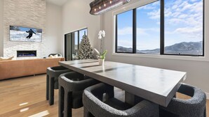 A modern dining area with a large table, dark chairs, a fireplace, a television, and large windows with a mountain view. - East Klaim Drive Departments (Hideout)